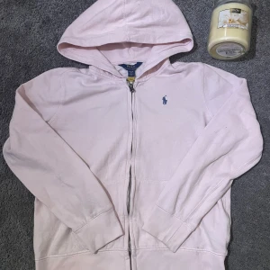 Ljusrosa hoodie Ralph Lauren - Säljer en ljusrosa hoodie från Ralph Lauren med dragkedja och det klassiska logotypbroderiet på bröstet. Tröjan har långa ärmar och är tillverkad i en mjuk bomullsblandning. Perfekt för en avslappnad stil. Storlek XL (16)
