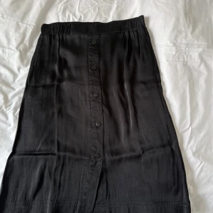 Svart kjol från House of Lola - Snygg svart kjol i glansigt tyg från House of Lola med dekorativa knappar framtill. Kjolen är tillverkad i en mjuk blandning av rayon och viskos, vilket ger den ett fint fall. Perfekt för en stilren look. Kjolen går strax nedsnför knäna och har en skön mjuk resår i midjan. Lätt att bara dra på sig.