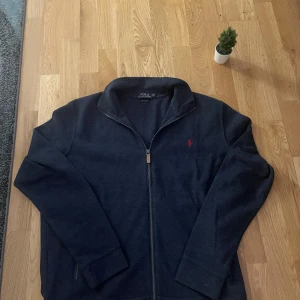 Mörkblå tröja med dragkedja från Ralph Lauren - Passar med storlek L möjlight viss M, men är XXL Säljer en stilren mörkblå tröja från Ralph Lauren med dragkedja och två praktiska fickor framtill. Tröjan har en liten röd logotyp på bröstet och är perfekt för en avslappnad look. Passar bra till både jeans och chinos.