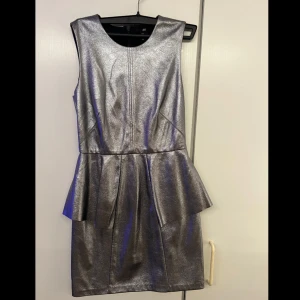 Silverfärgad klänning från H&M - Snygg silverfärgad klänning från H&M med en peplumdetalj i midjan. Klänningen är ärmlös och har en kort längd, perfekt för en festlig kväll. Materialet ger en glittrig look som verkligen sticker ut.