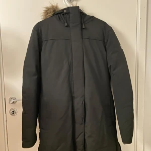 Vinterjacka  - Säljer en svart vinterjacka från Mountain Works med en mysig pälsfodrad huva. Jackan har långa ärmar och en stilren design, perfekt för kalla dagar. Den är utrustad med dragkedja och knappar för extra skydd mot kylan.