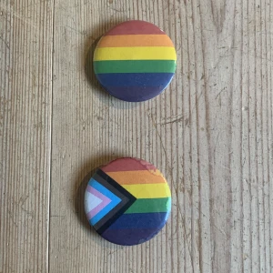 Regnbågsfärgade pins - Två färgglada pins med regnbågsdesign. Den ena har den klassiska regnbågsflaggan medan den andra har en progressiv design med extra färger för inkludering. Perfekt för att visa ditt stöd och sprida kärlek och acceptans. 🏳️‍🌈