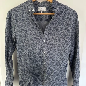 Mönstrad skjorta från Hampton Republic - Säljer en snygg mönstrad skjorta från Hampton Republic. Skjortan är i en grå nyans med ett coolt blommigt mönster. Den har långa ärmar och knappar framtill. Perfekt för både vardag och fest!