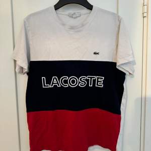 Säljer en snygg t-shirt från Lacoste i flera färger. Den har en vit överdel, en mörkblå mittendel med Lacoste-loggan i vitt, och en röd nederdel. T-shirten är kortärmad och perfekt för en casual look. 🐊