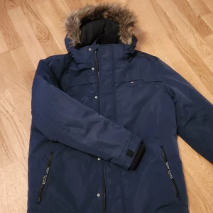 Blå vinterjacka från Tommy Hilfiger - Säljer en snygg och varm blå vinterjacka från Tommy Hilfiger. Jackan har en avtagbar huva med pälsimitation och är fodrad med tekniska fibrer för extra värme. Den har en dragkedja framtill och flera praktiska fickor. Perfekt för kalla vinterdagar!