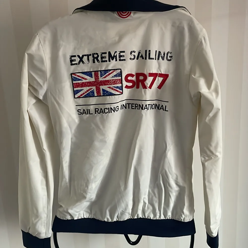 Säljer en snygg vit jacka från Sail Racing med SR77-tryck och brittisk flagga på ryggen. Jackan har dragkedja och två fickor med dragkedjor. Perfekt för vår och höst, med en sportig stil. Den är i ny och passar perfekt för segling eller andra utomhusaktiviteter.. Takit.