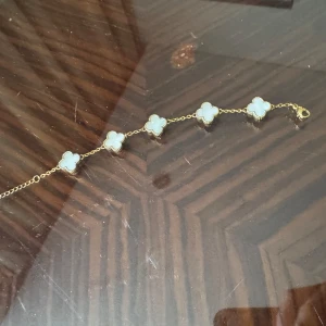 Guldarmband med fyrklöverdesign - Snyggt guldarmband med fem fyrklöverformade hängen i vit pärlemor. Perfekt för att ge en touch av elegans till vilken outfit som helst. Armbandet har en fin kedja och ett smidigt lås. Passar både till vardags och festliga tillfällen.