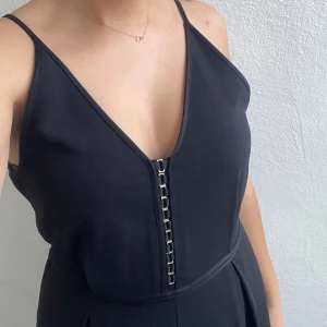 Svart  jumpsuit  - Säljer en svart jumpsuit med smala axelband och v-ringning. Klänningen har en guldiga knäppen framtill och är perfekt för festliga tillfällen. Tyvärr finns en liten skada på tyget, se bild, därav priset  