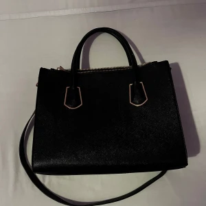 Svart läderväska väska, guld - Bra skick, hårt material. Guld detaljer och resten är svart. Handtagen står rakt up. Två stora fickor med dragskedja och en mindre med dragskedja, alla dragskedjor fungerar. Två mindre fickor på insidan 