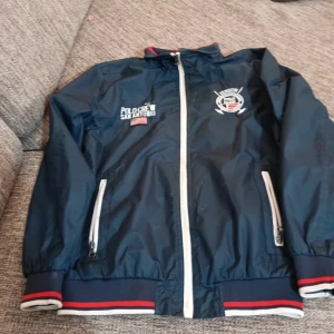 Mörkblå vindjacka från Vinson Polo Club - Säljer en snygg mörkblå vindjacka från Vinson Polo Club. Jackan har dragkedja fram och broderade detaljer med texten 'Polo Crew San Antonio'. Den har ribbade muddar med röd och vit rand. Perfekt för vår och höst!