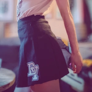 Svart kortkjol med tryck - Säljer en svart kortkjol med ett coolt tryck på sidan. Kjolen har en plisserad design som ger en snygg och ungdomlig look. Perfekt för både vardag och fest! 🖤