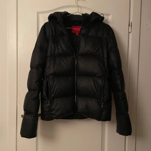 Svart pufferjacka från Guess - Säljer en snygg svart pufferjacka från Guess i storlek XS. Jackan har en innerficka och justerbar luva. Använd en vinter inga skador. Sitter även mer som en S/M.
