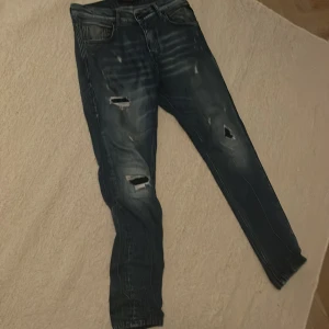 Blå jeans med slitningar - Säljer ett par snygga blå jeans från Replay med coola slitningar på benen. De har en slim passform och är perfekta för en avslappnad stil. Jeansen har en klassisk femficksdesign och knappar i midjan. Perfekta för både vardag och fest!