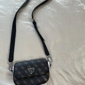Svart axelväska från Guess - Snygg svart axelväska från Guess. Väskan har en justerbar axelrem och en praktisk storlek för vardagsbruk. Perfekt för att ge din outfit en touch av lyx och stil.