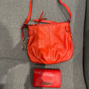  Orange axelväska och plånbok från DKNY - Snygg klar orange axelväska i skinn från DKNY med matchande plånbok. Väskan har en justerbar axelrem och en dekorativ metallbricka med DKNY-logga. Perfekt för att ge en färgklick till din outfit. Använd men i ok skick. Fler foton finns 😊.