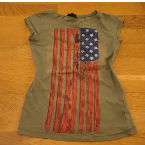 Grön t-shirt med flaggmönster från KappAhl - Säljer en grön t-shirt från KappAhl med USA tryck. T-shirten har korta ärmar och en rund halsringning