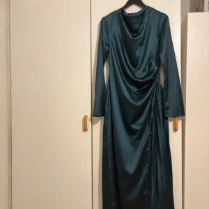 Mörkgrön långklänning i satin - Elegant mörkgrön långklänning i satin med draperad framsida och långa ärmar. Perfekt för en stilfull kväll.