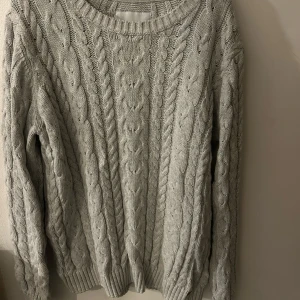 Grå stickad tröja - Mysig grå stickad tröja med klassiskt flätmönster. Perfekt för kyliga dagar med sin bekväma passform och långa ärmar. Ett måste i garderoben för en stilren look.