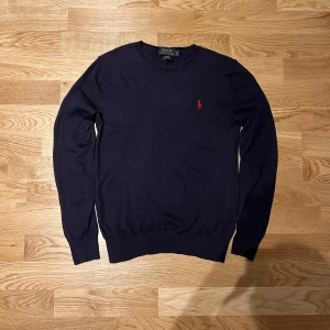 Mörkblå tröja från Polo Ralph Lauren - Snygg mörkblå tröja från Polo Ralph Lauren i merinoull. Tröjan har en slim fit passform i storlek S men passar som XS, och ett rött broderat logotypmärke på bröstet. Perfekt för en stilren look.