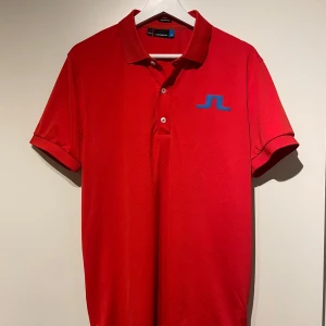 Golfpiké J.Lindeberg - Säljer en snygg röd/orange golfpiké från J.Lindeberg i storlek M. Tröjan har en klassisk krage och knappar framtill. Den är perfekt för både vardag och sport. Den blå loggan på bröstet ger en cool kontrast. 