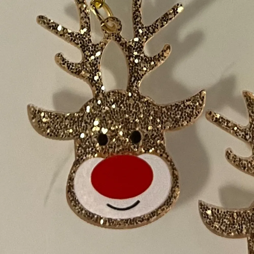 Charmiga örhängen i form av renar med glittrig yta och röd nos. Perfekta för att sprida julstämning med sin lekfulla design. De hängande örhängena har guldfärgade krokar och detaljer.. Asusteet.