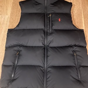 Svart dunväst från Ralph Lauren - Snygg svart dunväst från Ralph Lauren med dragkedja och ikonisk logga på bröstet. Perfekt för kyligare dagar och ger en stilren look. Västen har praktiska fickor med dragkedja. Skicket är 10/10 knappast använd, vid funderingar är de bara att fråga. Nypris är 2695kr mitt pris 1000kr