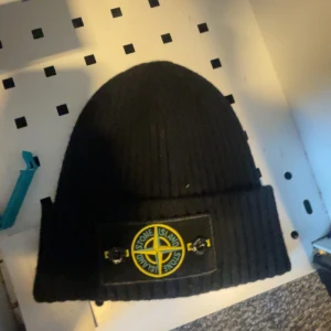 Svart ribbad mössa från Stone Island - Snygg svart ribbad mössa från Stone Island med det ikoniska märket framtill. Perfekt för att hålla värmen under kyliga dagar. En stilren accessoar för alla tillfällen.