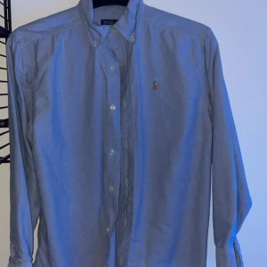 Blå skjorta från Polo Ralph Lauren - Säljer en klassisk blå skjorta från Polo Ralph Lauren med broderad logotyp på bröstet. Skjortan har långa ärmar och knappar framtill. Perfekt för en stilren look. Använd en gång på skolavslutningen! Skick 10/10🔥frågor om annat bara fråga