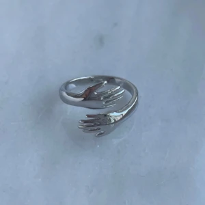 Harmony Hands Ring från Popy i silver - Justerbar silverring med en kreativ design av två händer som omsluter fingret. Perfekt för att ge en personlig touch till din stil. Kommer i en söt rosa påse.
