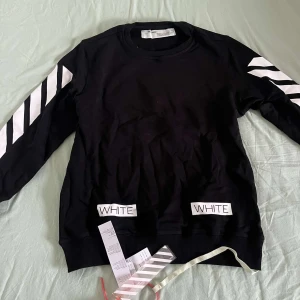 Svart tröja från Off-White - Snygg svart tröja från Off-White med vita diagonala ränder på ärmarna och textdetaljer fram och bak. Perfekt för en stilren look.
