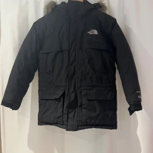Svart vinterjacka från The North Face 10-12 år - Svart barnparka (10-12år) från The North Face (Hyvent 550) med fakepäls på huvan. Vadderingen består av återvunnen dun och den avtagbara fuskpälskragen består av akryl och polyester. Yttermaterialet är vatten - och vindavvisande. Jackan är utrustad med en högre krage och en rejäl dragkedja samt kardborre. Fickor med lock och kardborre finns framtill och på bröstet finns fickor som stängs med dragkedja. Perfekt för kalla dagar. Använd av två barn men i bra skick! Namnmärkt.