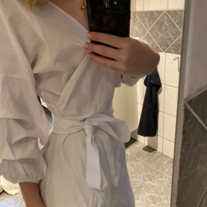 Vit omlottklänning med knytband - Säljer en stilren vit omlottklänning med knytband i midjan. Klänningen har långa ärmar och en v-ringad design. Perfekt för studenten eller till sommaren. Använd 2 gånger