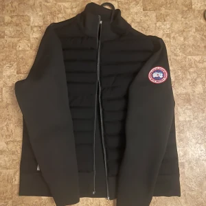 Svart canada goose jacka  - Snygg svart jacka från Canada Goose med ett tydligt emblem på ärmen. Jackan har en dragkedja framtill och är quiltad på framsidan. Perfekt för kyligare dagar.