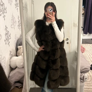 Brun lång pälsväst - Snygg och elegant brun lång pälsväst perfekt för kyliga dagar. Västen har en lyxig look med sitt fluffiga material och är utan ärmar, vilket gör den lätt att bära över en tröja eller blus.