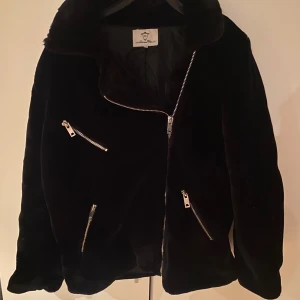 Svart fuskpälsjacka  - Snygg svart fuskpälsjacka med dragkedjor. Jackan har en cool bikerstil med asymmetrisk dragkedja och två snedställda fickor med dragkedjor. Perfekt för att hålla sig varm och stilren.