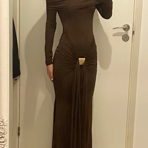 Brun långklänning med drapering - Elegant brun långklänning med offshoulder-design och drapering framtill. Klänningen har långa ärmar och en tight passform som framhäver figuren. Perfekt för en stilfull kväll. Riktiga färgen som inte framgår tydligt i bilden är brun/mörk vinröd