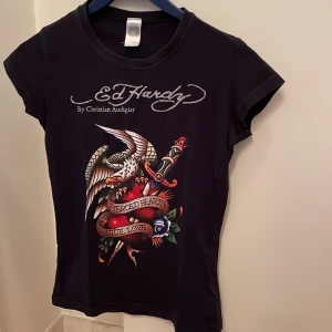 Svart Ed Hardy t-shirt med tryck - Snygg svart t-shirt från Ed Hardy med ett stort färgglatt tryck av en örn och ett hjärta med texten 'True Love'. T-shirten har korta ärmar och rund halsringning. Perfekt för en cool och avslappnad stil.
