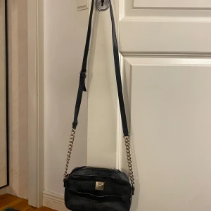 Svart axelväska med kedjedetaljer - Snygg svart axelväska från Guess med kedjedetaljer och ett diskret mönster. Väskan har en dragkedja upptill och en liten metallplatta med logotyp på framsidan. Perfekt för att bära dina nödvändigheter med stil. Defekter: Rost på kedjedetaljerna och märket samt lite sliten. 