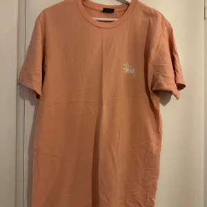 Persikofärgad t-shirt från Stussy - Säljer en snygg persikofärgad t-shirt från Stussy med deras ikoniska logga tryckt på bröstet. T-shirten är kortärmad och har en klassisk rund halsringning. Perfekt för en avslappnad stil.