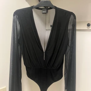 Svart djup v-ringad bodysuit - Snygg svart bodysuit från Gina med djup v-ringning och transparenta meshärmar. Perfekt för en kväll ute eller en speciell tillställning. Den har en åtsittande passform och är tillverkad i ett mjukt och stretchigt material. Mycket bra skick!