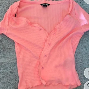 Rosa ribbad topp från Monki - Säljer en söt rosa ribbad topp från Monki med knappar framtill. Toppen har korta ärmar och en lätt vågig kant vid ärmslut och nederkant. Perfekt för en avslappnad stil.