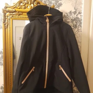 Svart jacka från Michael Kors  - Snygg svart jacka från Michael Kors med dragkedja och huva. Jackan har långa ärmar och praktiska fickor med dragkedjor. Perfekt för kyligare dagar. Windbreaker med gosigt foder 