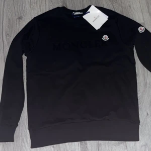 Svart tröja från Moncler - Snygg svart tröja från Moncler med deras logga på bröstet och ärmen. Tröjan har långa ärmar och en klassisk passform. Perfekt för en stilren look. Dessutom är den aldrig använd med alla tags kvar. 