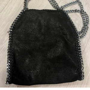 Svart axelväska med kedjedetaljer - Snygg svart axelväska med kedjedetaljer längs kanterna. Väskan har en glittrig yta som ger den en extra touch av stil. Perfekt för att lyfta vilken outfit som helst.