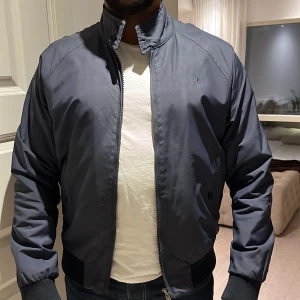 Mörkblå bomberjacka - Snygg mörkblå bomberjacka med dragkedja och ribbade muddar. Jackan har en stilren design med en liten broderad logga på bröstet. Perfekt för en casual look.