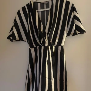 Svartvit randig klänning - Snygg svartvit randig klänning med v-ringning och korta ärmar. Perfekt för en stilren look. Klänningen har en omlott-design som ger en smickrande passform. Silke liknande tyg.