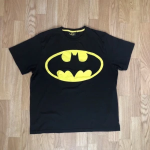 Svart Batman Printed T-Shirt  - Cool svart t-shirt med det ikoniska Batman-logotypen i gult på bröstet. Perfekt för alla superhjältefans! T-shirten har en rund halsringning med gul kant och korta ärmar.