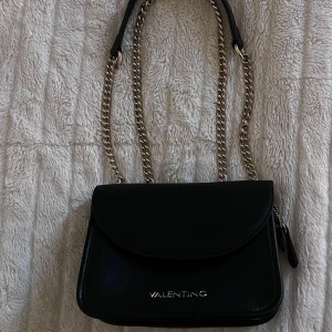 Svart axelväska från Valentino - Elegant svart axelväska från Valentino med kedjedetaljer och dragkedja. Perfekt för att bära dina nödvändigheter med stil. Väskan har en stilren design med ett lock och en säker stängning.
