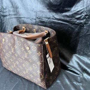 Brun monogram handväska - Elegant brun handväska med klassiskt Louis Vuitton monogrammönster. Väskan har två handtag och detaljer i läder. Perfekt för en stilren look.