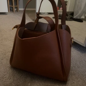 Brun handväska med dragkedja - Snygg brun handväska i skinn med praktisk dragkedja. Perfekt för att bära dina dagliga nödvändigheter med stil. Väskan har en elegant design med två handtag och en justerbar axelrem.